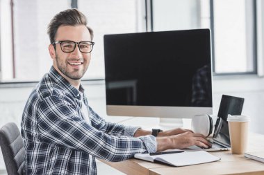 handsome young programmer in eyeglasses smiling at camera while working with laptop and desktop computer clipart