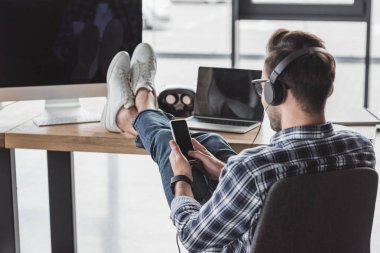 dizüstü bilgisayar ve masaüstü bilgisayar ile masada bacakları ile otururken smartphone kullanarak Kulaklığımda genç adam