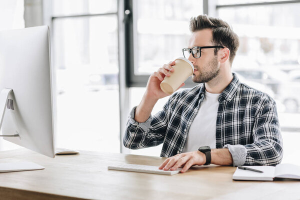 young man in eyeglasses drinking coffee from paper cup and using desktop computer