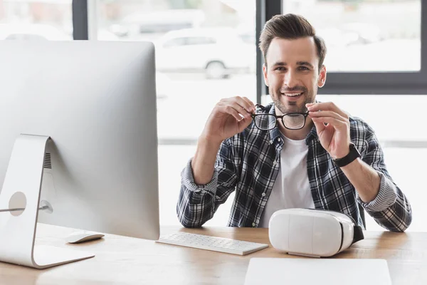 Guapo Joven Programador Sosteniendo Anteojos Sonriendo Cámara Mientras Está Sentado — Foto de Stock