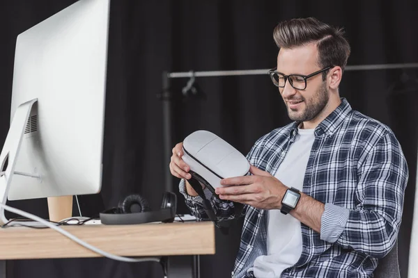 smiling young man in eyeglasses holding virtual reality headset at workplace