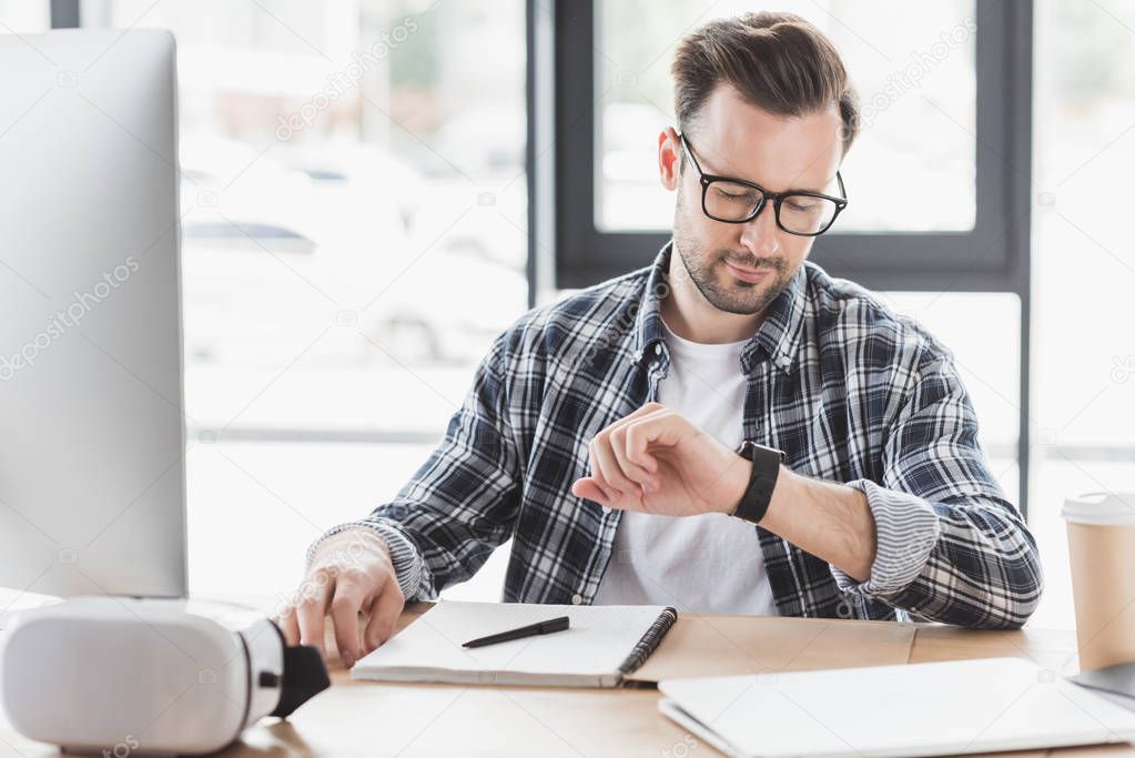 young man in eyeglasses checking smartwatch while sitting at workplace