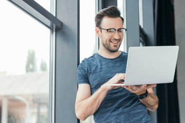 smiling young man in eyeglasses using laptop near window clipart