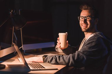 dizüstü ve masaüstü bilgisayarla çalışırken gitmek için gözlük kahve tutan genç programcı gülümseyen