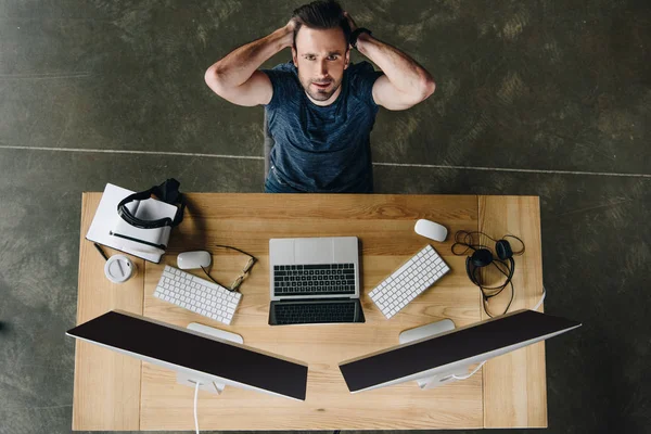 handsome young man with hands behind head looking at camera while sitting at workplace with computers