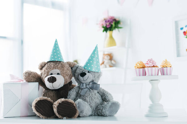 плюшевые мишки в конусах на столе с подарочной коробкой и кексами
 