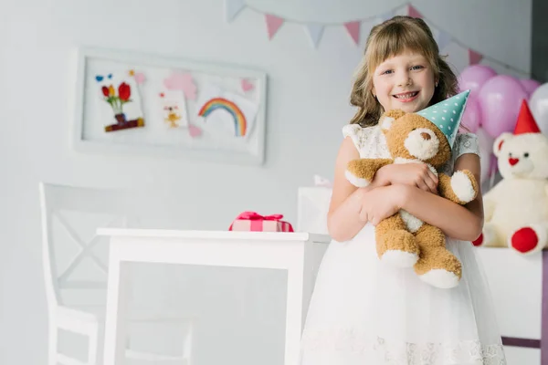 adorable birthday kid looking at camera and holding teddy bear in cone
