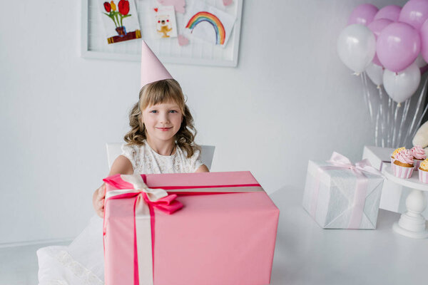 smiling birthday kid in cone sitting at table with huge gift box 