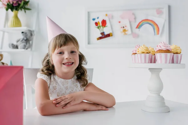 Lindo Cumpleaños Niño Cono Mirando Cámara Sentado Mesa Con Cupcakes — Foto de stock gratis