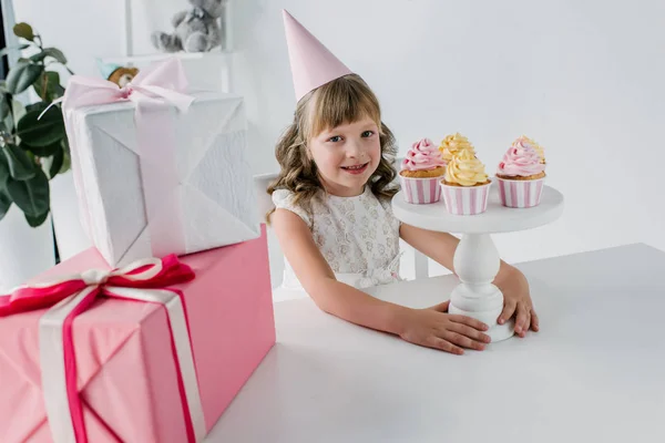 Lächelndes Geburtstagskind Kegel Sitzt Tisch Mit Cupcakes Und Geschenkschachteln — kostenloses Stockfoto