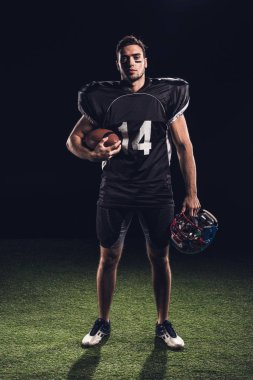 confident american football player in black uniform holding helmet and ball and looking at camera on black