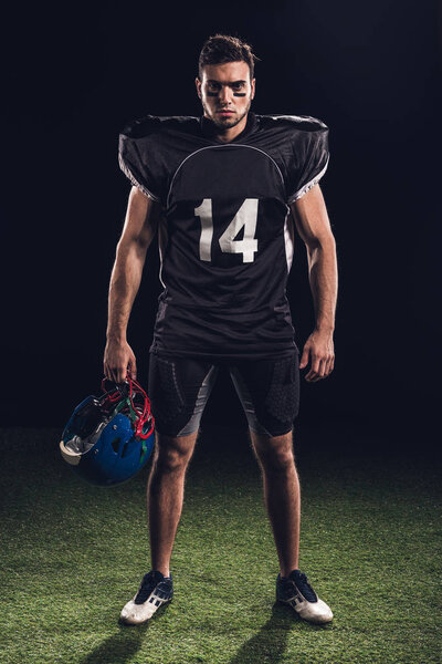 confident american football player in black uniform holding helmet and looking at camera on black