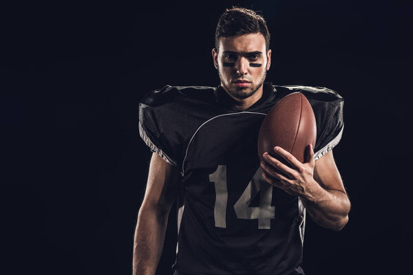 young serious american football player with ball looking at camera isolated on black