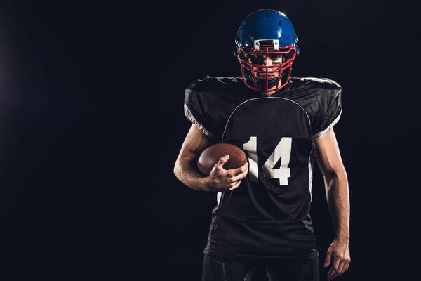 confident american football player holding ball in hand and looking at camera on black