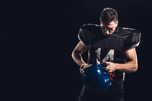 american football player in black uniform with helmet in hands isolated on black