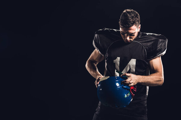 american football player in black uniform with helmet in hands isolated on black