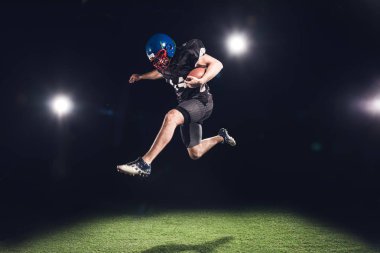 american football player jumping with ball over green grass under spotlights on black clipart