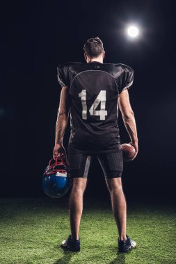 rear view of american football player with ball and helmet standing on grass under spotlight on black