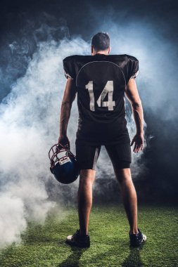 rear view of equipped american football player with helmet in hand against white smoke clipart