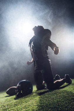 young depressed american football player looking up and shouting while standing on knees on green grass against white smoke clipart