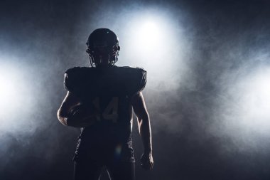 dark silhouette of equipped american football player with ball against white smoke clipart