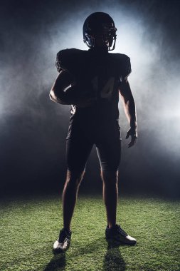 silhouette of equipped american football player with ball standing on green grass against white smoke clipart