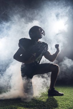 yeşil çimenlerin üzerinde diz üzerinde duran ve beyaz duman içinde yumruk yükselterek genç Amerikan futbolu oyuncusu