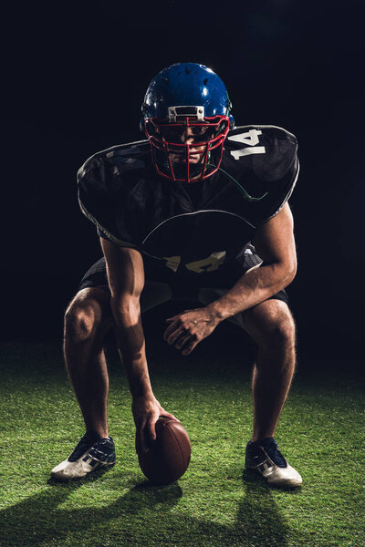 serious american football player standing on grass with ball and looking at camera on black