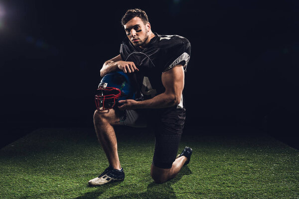 american football player standing on one knee on grass with helmet on black