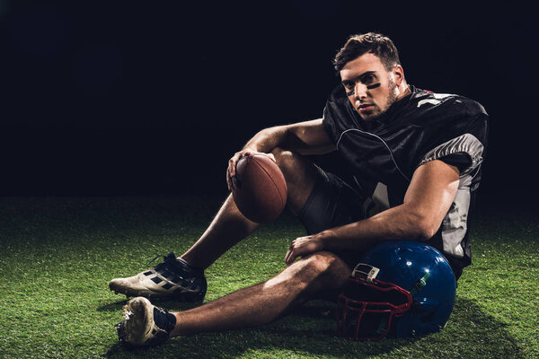 american football player sitting on grass with ball and helmet on black
