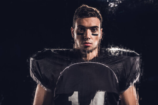 view of handsome american football player looking at camera on black through wet glass