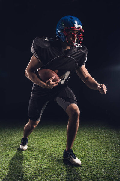 american football player holding ball and running on field on black