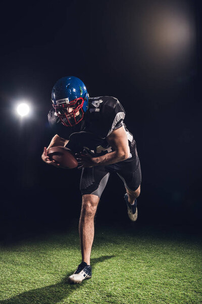 american football player holding ball and running on field under spotlights on black