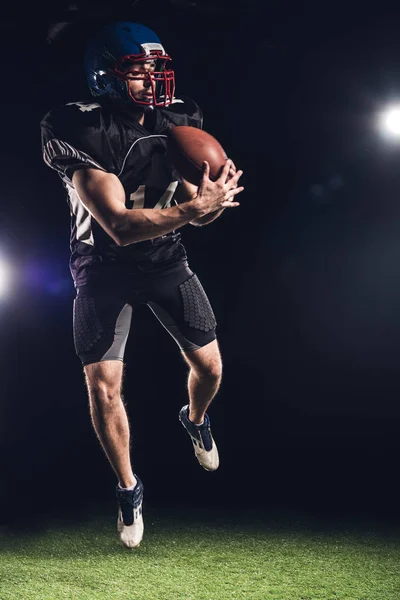 ブラック スポット ライトの下で緑の草の上のボールでジャンプ若いアメリカン フットボール プレーヤー — ストック写真