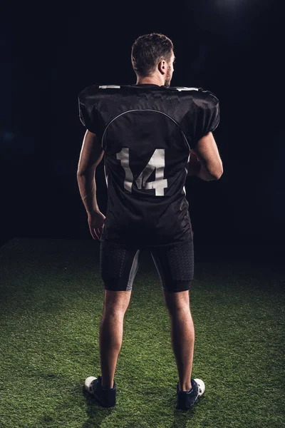 rear view of american football player in black uniform standing on grass on black