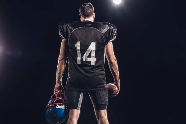黒のボールとスポット ライトの下のヘルメット アメリカン フットボール プレーヤーの背面図  — 無料ストックフォト