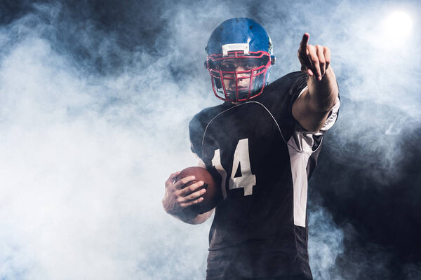 handsome american football player with ball pointing somewhere against white smoke