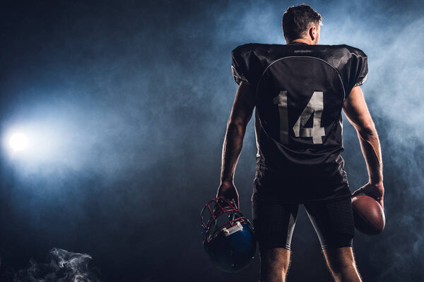 rear view of equipped american football player with helmet and ball in hands against white smoke