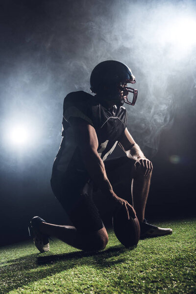 thoughtful american football player with ball standing on knee against white smoke