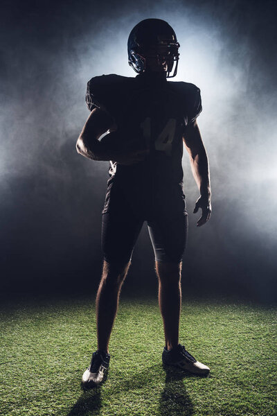 silhouette of equipped american football player with ball standing on green grass against white smoke