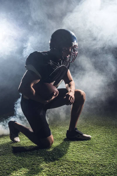 白い煙に対して手にボールで緑の草に膝の上に立って若いアメリカン フットボール プレーヤー — ストック写真
