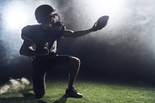 緑の芝生に膝の上に立っていると煙の白に対してボールを保持している アメリカン フットボール プレーヤー — ストック写真