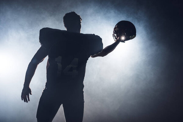 silhouette of american football player holding helmet against white smoke