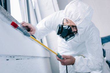 pest control worker in respirator spraying pesticides under windowsill at home clipart