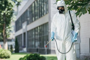 pest control worker in uniform and respirator spraying pesticides on street with sprayer   clipart