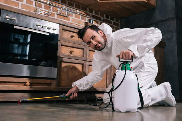 pest control worker spraying pesticides under cabinet in kitchen and looking at camera