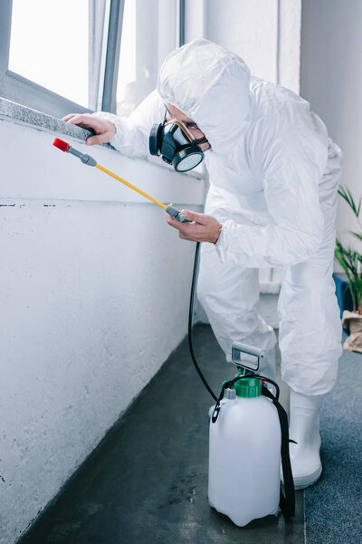 pest control worker in uniform spraying pesticides under windowsill at home