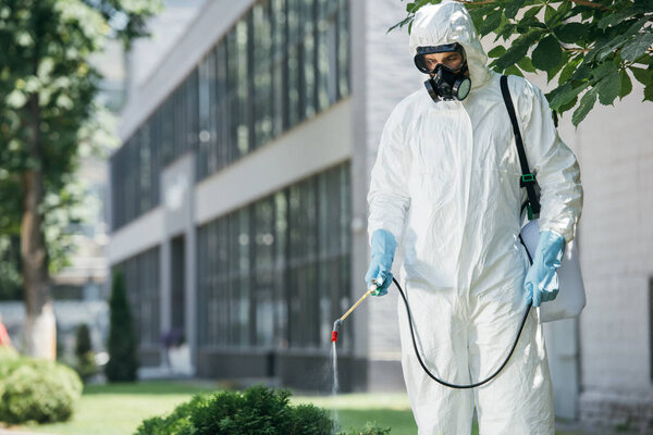 работник по борьбе с вредителями в форме и респираторе, распыляющий пестициды на улице с распылителем
  