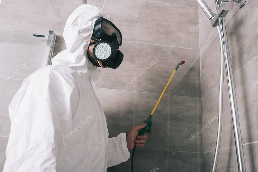 pest control worker in respirator spraying pesticides with sprayer in bathroom