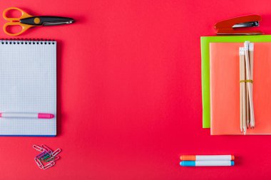 top view of arranged textbooks, stapler, markers, paper clips, scissors and blank notebook on red background  clipart